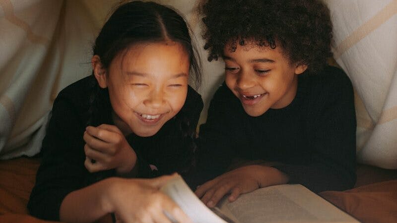 A Young Girl and a Boy Reading a Book while Under the Blanket