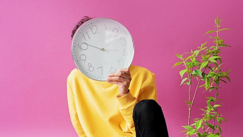 A Person in Yellow Sweater Sitting on the Floor Near the Potted Plants while Covering Face Using a Clock