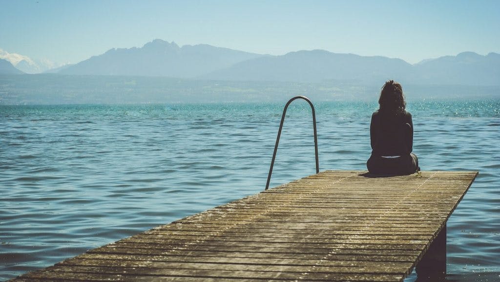 a woman sits on the end of a dock during daytime staring across a lake